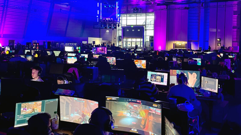 A wide shot of a bustling LAN party event, likely Assembly in Helsinki, filled with many individuals focused on their computers playing video games. The large hall is illuminated with vibrant blue and purple lighting, showcasing the energetic atmosphere of a major esports and gaming gathering that Hive Helsinki students often attend.