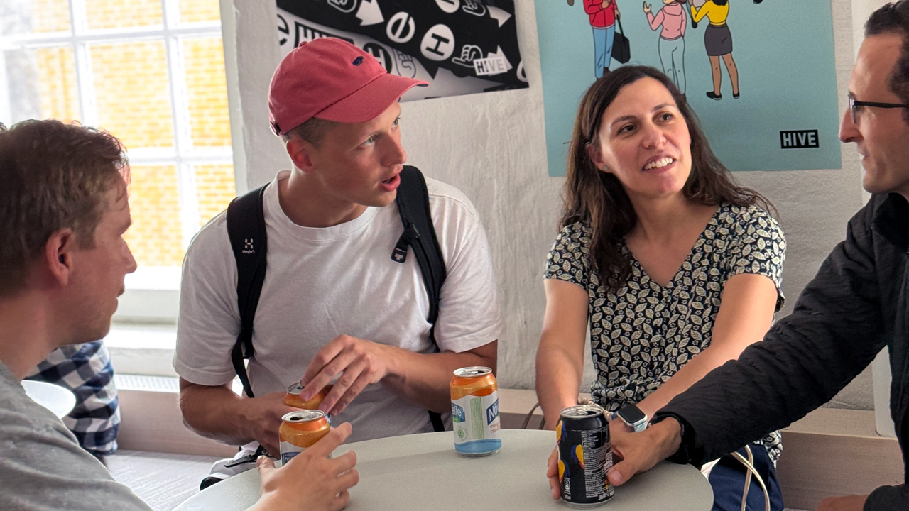 A group of Hive Helsinki students or community members having a casual conversation around a table, holding drinks. The person in the red cap is speaking animatedly. In the background, posters with "HIVE" branding are visible, representing the relaxed and social atmosphere at the tech education campus in Helsinki, Finland.