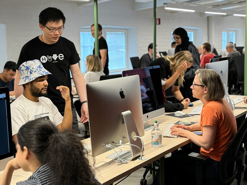 Students at Hive Helsinki's modern coding lab, actively engaged with Apple iMac computers. One student wearing a Hive t-shirt is helping another, showcasing the peer-to-peer learning and collaborative environment at the tech education campus in Helsinki, Finland.