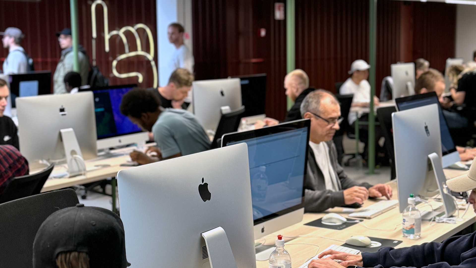 An active computer lab at Hive Helsinki, featuring rows of students intensely focused on Apple iMac computers. Several individuals are visible, typing and working at their desks. In the background, a large neon hand sign is displayed on a dark red textured wall.
