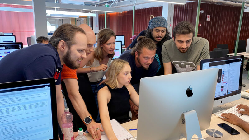 A group of Hive Helsinki students gathered around an Apple iMac, collaboratively working on a coding project. One student is actively typing, while others lean in to observe the screen, demonstrating the hands-on, peer-to-peer learning approach fostered at the coding school in Helsinki, Finland.