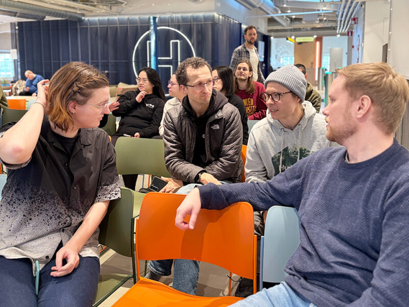 A group of students engaged in conversation and listening during an event at Hive Helsinki. They are seated on a mix of orange and green chairs, with some looking towards each other in discussion. In the background, a large 'H' logo is visible on a dark wall, and other attendees are blurred in the distance.