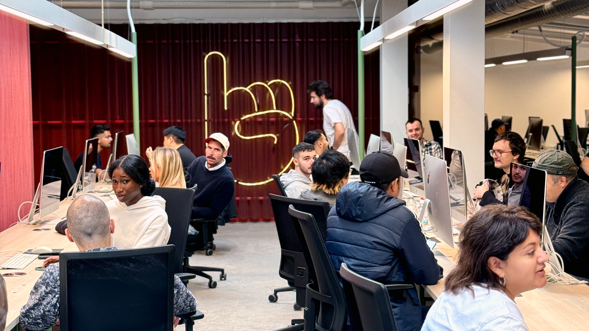 An active open-plan workspace at Hive Helsinki, filled with people working on Apple iMac computers. Many individuals are focused on their screens, while some are interacting or looking around. A prominent neon hand sign is visible on a red textured wall in the background, adding a modern, vibrant touch to the collaborative environment.
