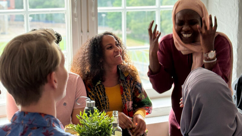 A group of diverse Hive Helsinki students laughing and socializing by a large window, enjoying a casual moment on campus. This image captures the vibrant and friendly atmosphere, promoting the strong community and peer interaction that defines the coding school experience in Helsinki, Finland.