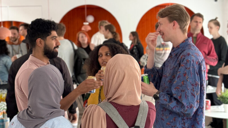 A diverse group of Hive Helsinki students and community members socializing at an event, with some holding drinks. The modern interior features distinctive orange arched wall sections in the background, highlighting the vibrant and inclusive atmosphere of the tech education campus in Helsinki, Finland.