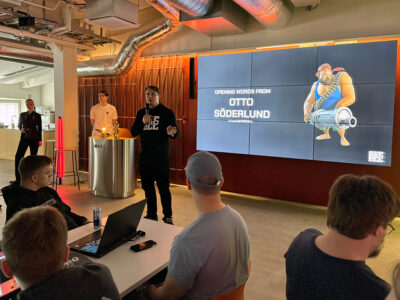 A presentation taking place at Hive Helsinki, with a speaker standing in front of a large screen displaying "OPENING WORDS FROM OTTO SÖDERLUND" and an illustration of a cartoon character. An audience is seated, facing the speaker, with some people working on laptops. The space has an industrial design with exposed pipes on the ceiling and warm lighting.