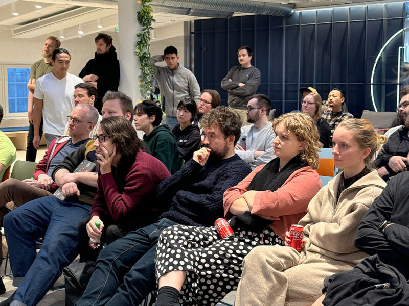 A diverse audience at the Supercell Challenge 2024 event at Hive Helsinki, attentively listening to a presentation. Individuals are seated and engaged, some holding drinks. This image captures the vibrant community and interest in coding challenges and tech careers at the Hive Helsinki campus in Finland.