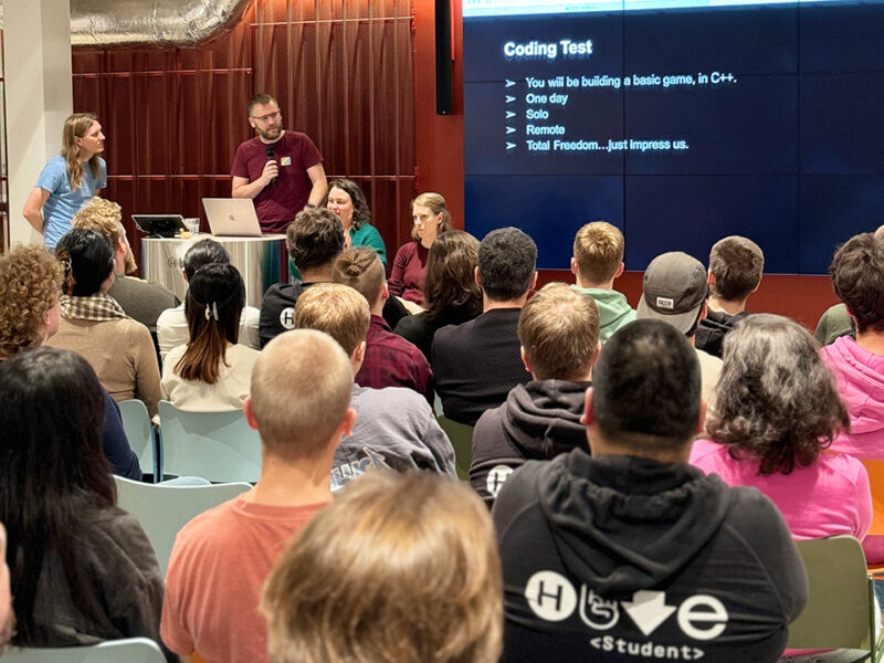 A speaker at the Supercell Challenge 2024 at Hive Helsinki explains the "Coding Test" rules to an audience. The screen lists details like "You will be building a basic game in C++," "One day," and "Total Freedom...just impress us." This image highlights the practical coding challenges and skills-based learning approach at Hive Helsinki, preparing students for tech careers in Finland.