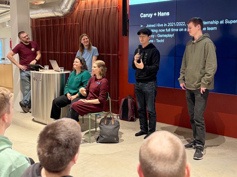 Presenters on stage at the Supercell Challenge 2024 at Hive Helsinki, introducing the event. A large screen displays names "Caruy + Hans" and details about internships at Supercell. This scene emphasizes the career pathways and industry connections available through Hive Helsinki's coding education in Finland.