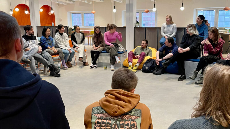 A diverse group of Hive Helsinki students engaged in a relaxed, circular discussion during the Winter Piscine 2024. They are seated on beanbags and chairs in a modern lounge area, highlighting the collaborative and community-focused learning environment at our Helsinki campus in Finland.