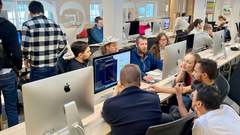 ive Helsinki students gathered around Apple iMac computers during the Winter Piscine 2024, actively engaged in peer-to-peer learning and problem-solving. Some individuals are leaning in to observe a screen, demonstrating the collaborative and hands-on nature of our coding education in Helsinki, Finland.