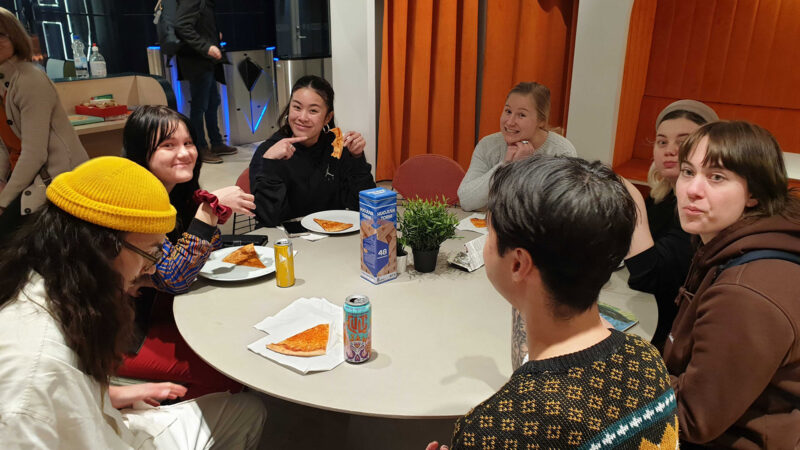 A small group of Hive Helsinki students enjoying pizza and drinks around a table during a social break at the Winter Piscine 2024. They are smiling and interacting, showcasing the friendly and inclusive community atmosphere that complements the intensive coding education in Helsinki, Finland.