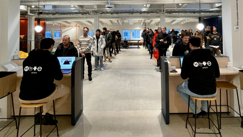 A long line of enthusiastic prospective students waiting to register for the Hive Winter Piscine 2024 in Helsinki, Finland. Two Hive staff members are at the check-in desks, assisting participants with laptops. This image captures the excitement and high demand for our immersive coding education program.