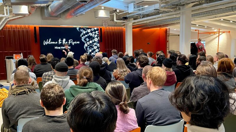A wide shot of an audience at the Hive Winter Piscine 2024 in Helsinki, attentively listening to a speaker on stage. The large screen behind the presenter displays "Welcome to the Piscine" and a DNA-like graphic. This image captures the engaging start of our intensive coding program in Finland.