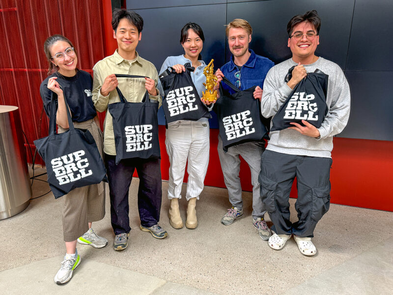 A group of five Hive Helsinki students, the winners of the Supercell AI Hackathon event, posing together. They are holding black "SUPERCELL" branded tote bags, and one person in the center is holding a golden trophy. The background features a dark wall and some red accents, suggesting a modern event space.