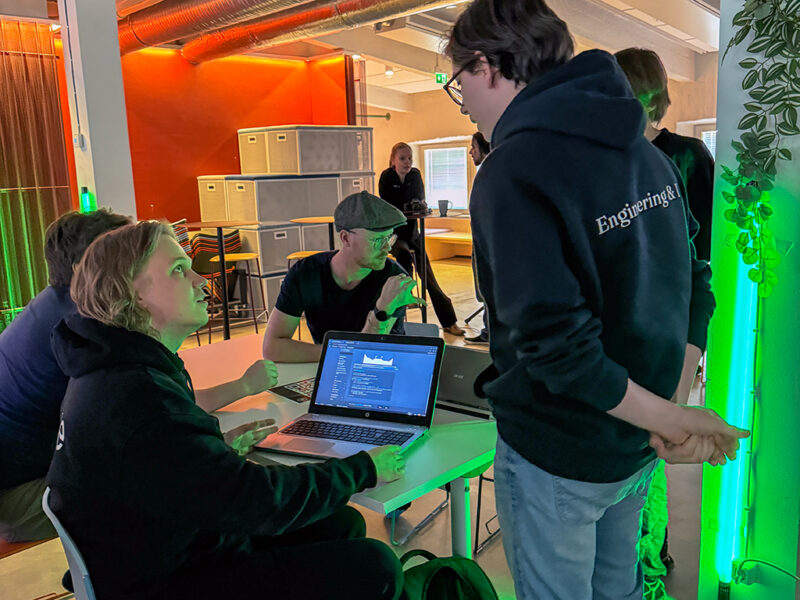 A group of students or individuals collaborating at the Supercell AI Hackathon at Hive Helsinki. One person is actively engaged with a laptop displaying data or code, while others are observing and discussing. The room features industrial elements, such as exposed ductwork, and some green lighting accents, suggesting a dynamic and creative environment.