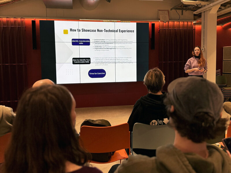 A presenter stands next to a large screen displaying slides titled "How to Showcase Non-Technical Experience" and "How to Identify Your Transferable Skills." The slides list bullet points related to communication, problem-solving, and organization. An audience is seated in front of the screen, facing away from the camera, listening to the presentation in a modern event space at Hive Helsinki.