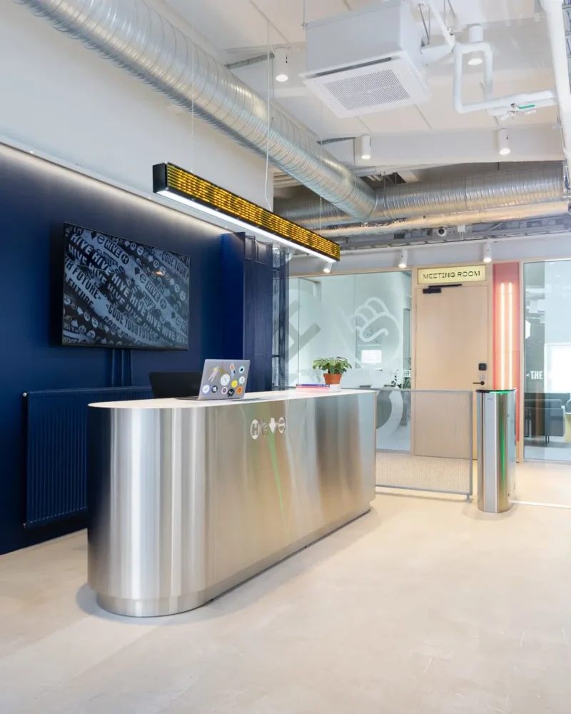 Modern reception area at Hive Helsinki with a sleek metallic front desk, a digital ticker display, and a meeting room entrance in a tech-inspired open office environment.
