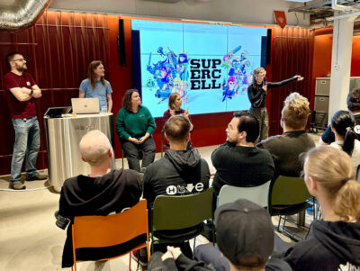 A speaker presents the Supercell Challenge 2024 to a seated audience at Hive Helsinki. The large screen behind them displays the Supercell logo and character art. This event highlights collaboration between Hive Helsinki and industry partners, offering real-world coding challenges and career opportunities in Finland's tech sector.