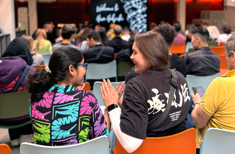 Two students, seen from behind, are seated in a lecture hall or event space, smiling and engaged in conversation. One student wears a brightly colored shirt with a tropical pattern, and the other wears a dark t-shirt. Many other attendees are visible in the background, facing a stage with a screen displaying text.