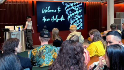 A group of students at Hive Helsinki during a welcome presentation. A speaker stands near a screen that says Welcome to Hive Helsinki. The atmosphere is friendly and creative, showing the inclusive community at the coding school in Finland.
