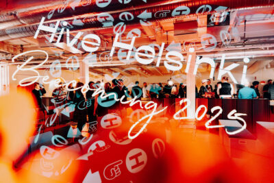 A wide view of people mingling at a Hive event in Helsinki, with warm orange tones, Hive branding, and “Hive Helsinki Becoming 2025” visible across the image.