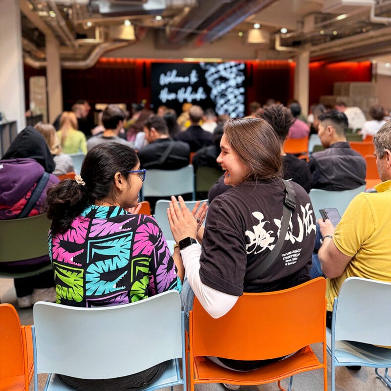 Two students, seen from behind, are seated in a lecture hall or event space, smiling and engaged in conversation. One student wears a brightly colored shirt with a tropical pattern, and the other wears a dark t-shirt. Many other attendees are visible in the background, facing a stage with a screen displaying text.