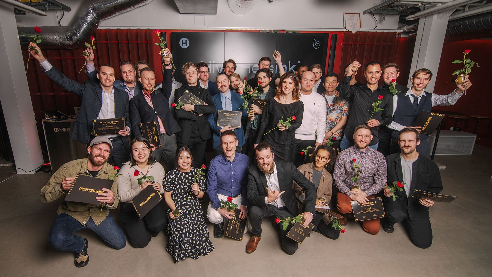A large group of smiling Hive Helsinki graduates posing together indoors. Many are holding diplomas or certificates and red roses, celebrating an achievement. Some individuals are standing, while others are kneeling in the front row. The background features exposed industrial ceilings and a dark wall with some text.