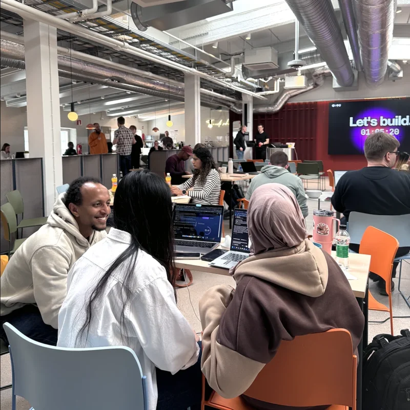 Three Hive students sitting around a table with laptops, turning toward the camera mid-conversation. Other students work and chat in the open campus space behind them, with a screen reading "Let's build" on the back wall.