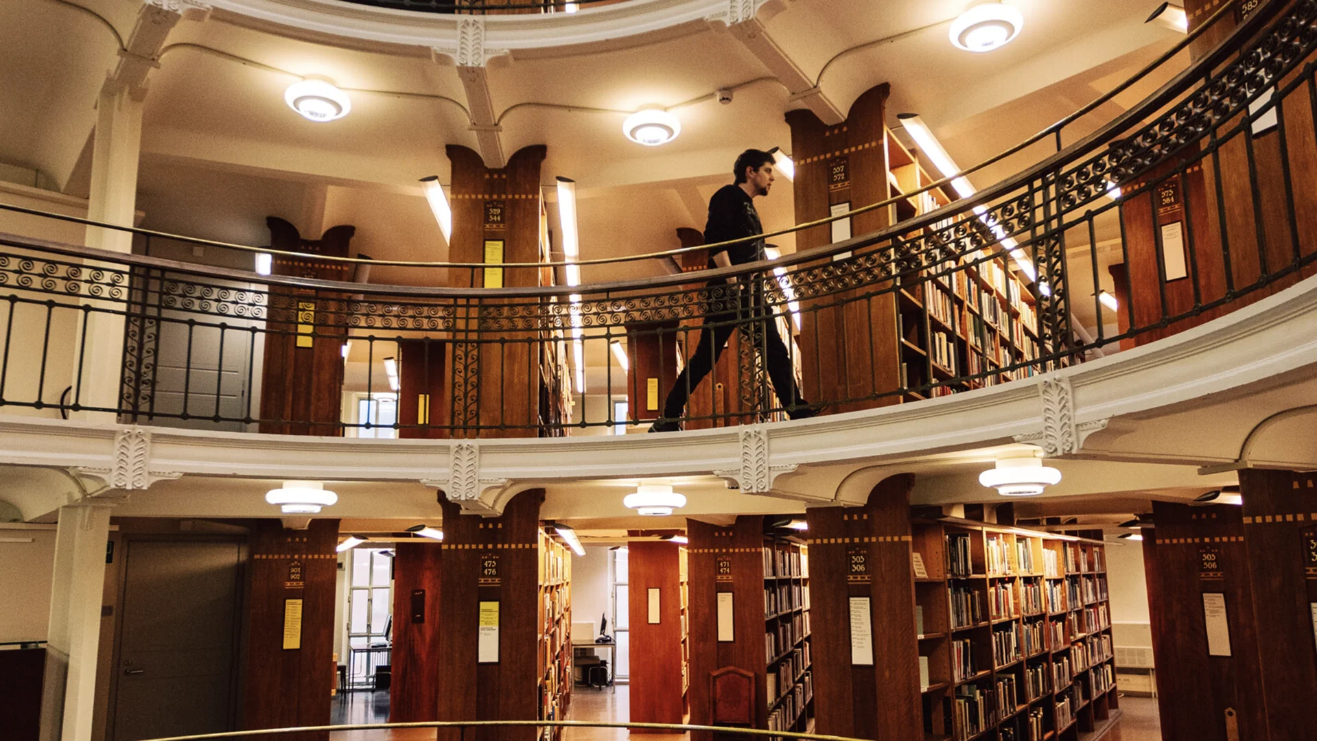Interior of the historic Rikhardinkatu Library in Helsinki: a warm-lit circular atrium with wooden bookshelves on the lower level, an ornate iron-railed gallery above, and a person walking along the upper balcony.
