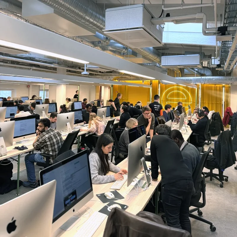 Open-plan tech workspace filled with rows of desktop computers and many people seated and standing working and collaborating, with exposed ceiling ducts, bright overhead lighting and a yellow glass partition wall with an arch design in the background.