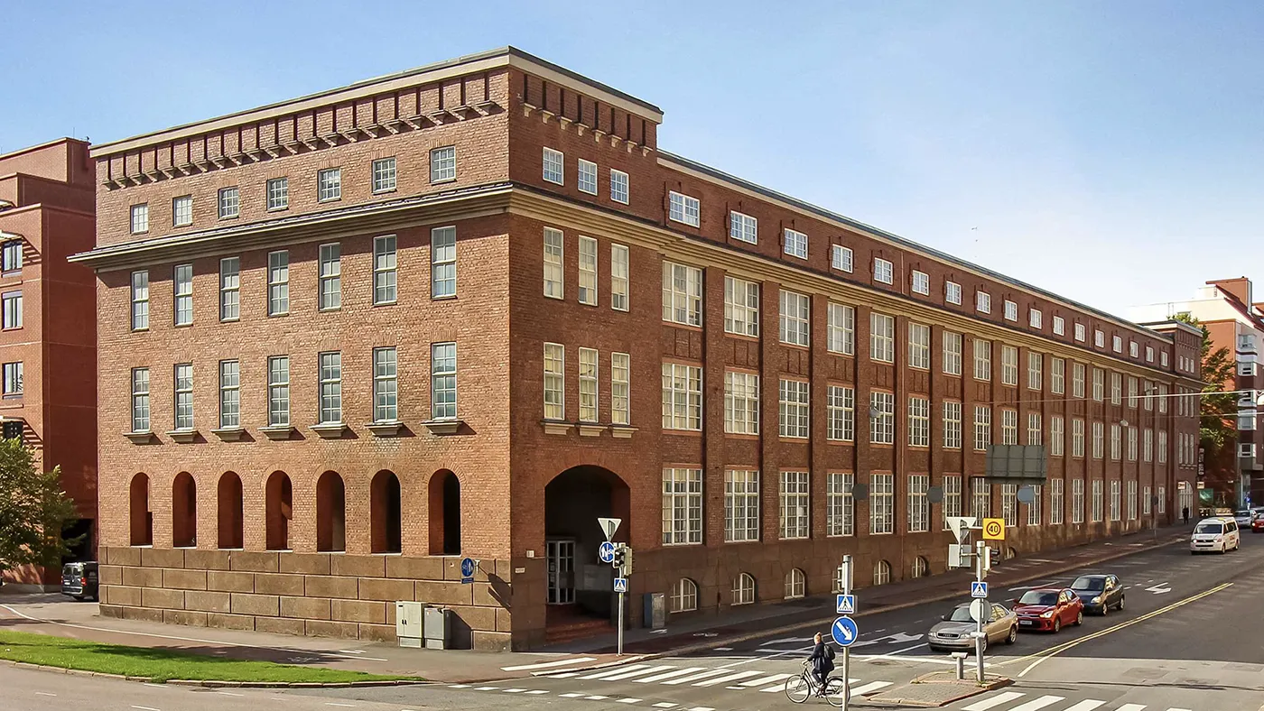 Exterior view of the Hive Helsinki campus building — a long red-brick, multi-story rectangular structure with arched openings on the ground floor and regular rows of windows, seen from a street corner with a crosswalk, parked cars and clear blue sky.