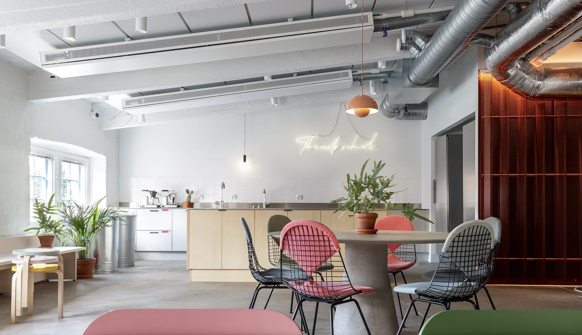 Bright, airy communal kitchen and lounge with a light-wood island, round dining table surrounded by mismatched wire-frame chairs with colorful cushions, potted plants, pendant lighting and a neon wall sign, large windows and exposed ceiling ducts with a red shelving accent.