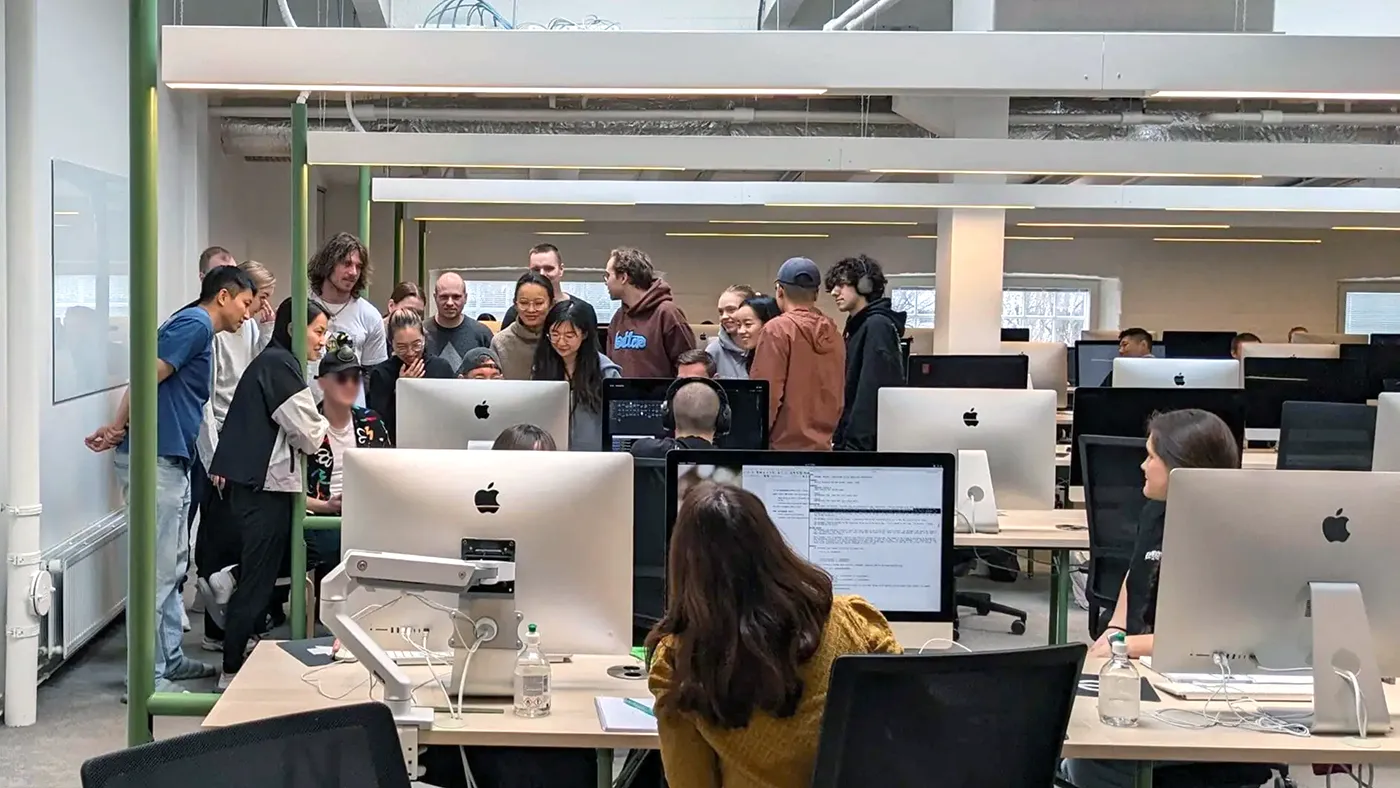 Group of students gathered around a computer screen in a bright, open-plan computer lab with rows of iMacs, collaborating on a project.