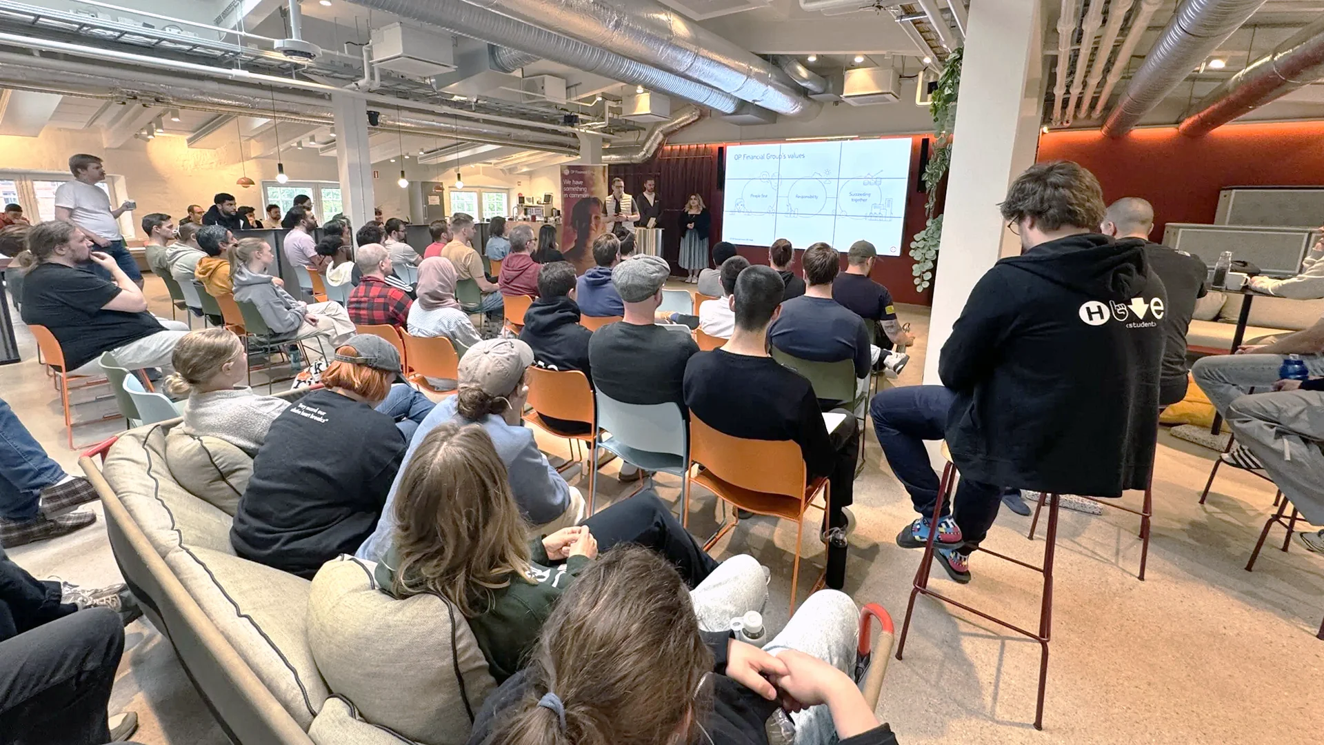 Students at an event in Hive Helsinki: a large audience seated on chairs and sofas in a modern open space with exposed ductwork, facing projection screens at the front.