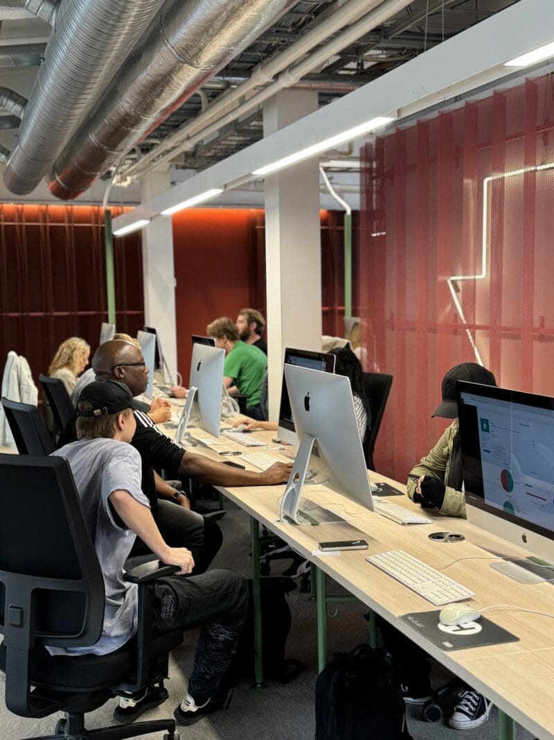 Open-plan office scene showing a long shared desk lined with iMacs and keyboards where several people in casual clothing sit and work — some wearing headphones — with exposed ductwork overhead, warm strip lighting and a red translucent partition in the background.