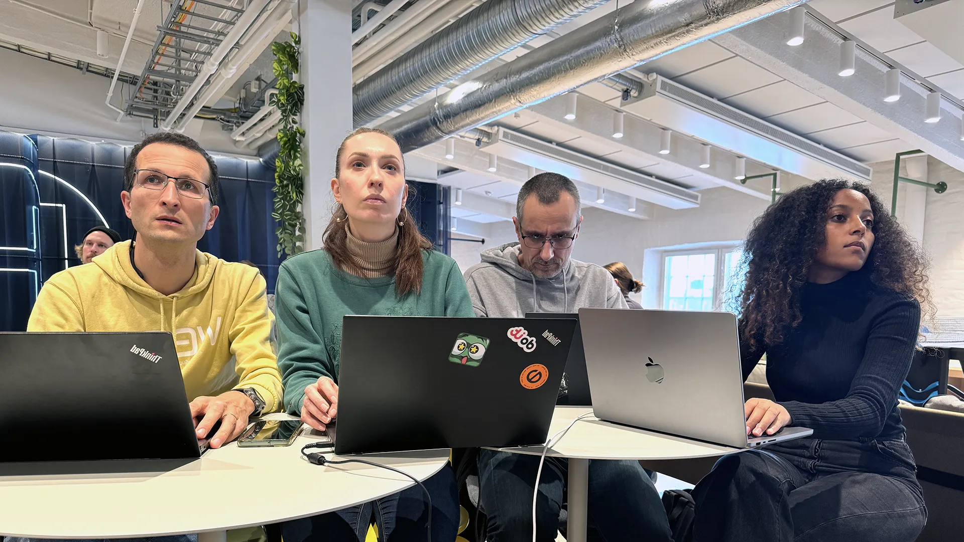 Four students seated side-by-side at a curved table working on laptops in a bright, modern open workspace with exposed ducts and industrial lighting; they appear focused and attentive, one laptop decorated with stickers.
