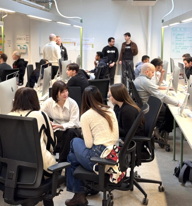 Open-plan coworking space with about a dozen adults seated at rows of desktop computers and clustered in small groups talking and collaborating, whiteboards and notes on the walls.
