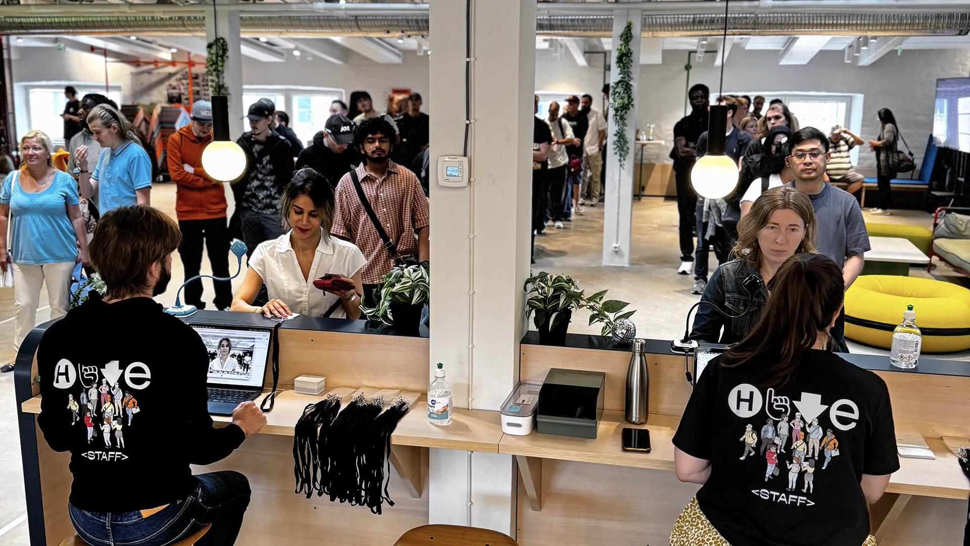 Busy indoor event registration area showing two staff in matching black T-shirts with a white graphic seated at a long wooden counter using laptops and handing out lanyards and materials while a long line of people waits behind them in an open-plan office with exposed beams, potted plants and yellow sofas.