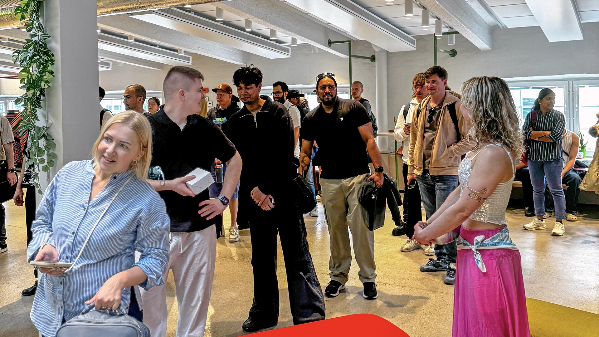 Bright indoor event space with a group of people standing and talking; a smiling woman in a light blue top sits in a wheelchair at left while others hold coats and bags, including a woman in a pink skirt at right.