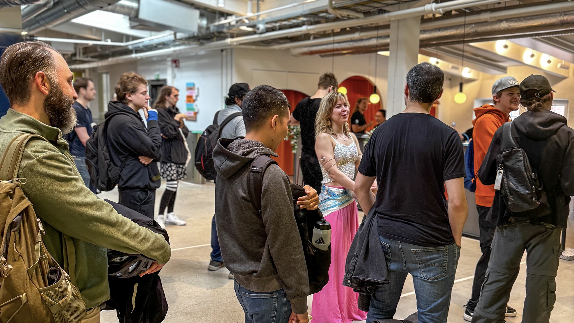 A group of people in casual clothing gathered inside an industrial-style event space with exposed ceilings; a woman in a bright pink and white dress stands near the center while others form a loose line or cluster.