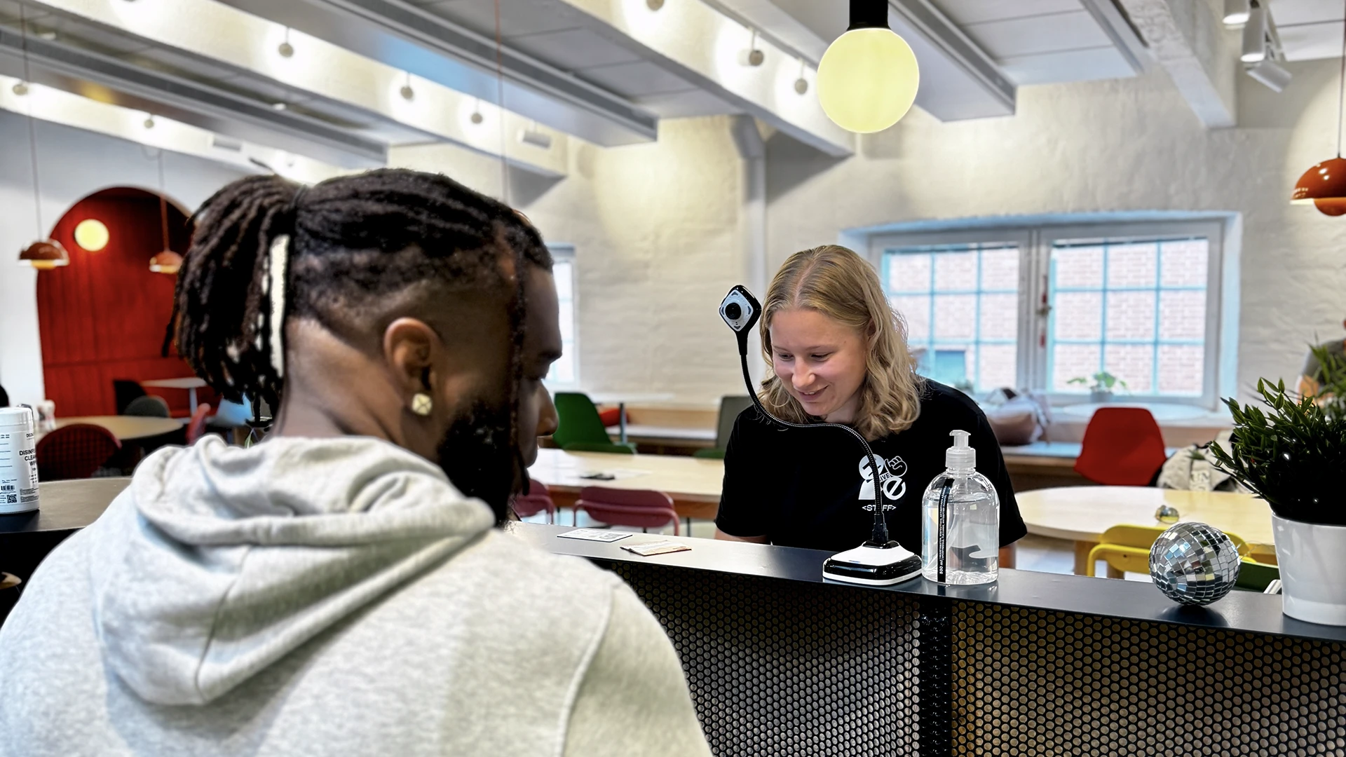 Person with braided hair seen from behind stands at a reception desk where a smiling receptionist with light hair engages with them in a bright, modern co‑working space with pendant lights overhead, a window in the background, and hand sanitizer and a potted plant on the counter.