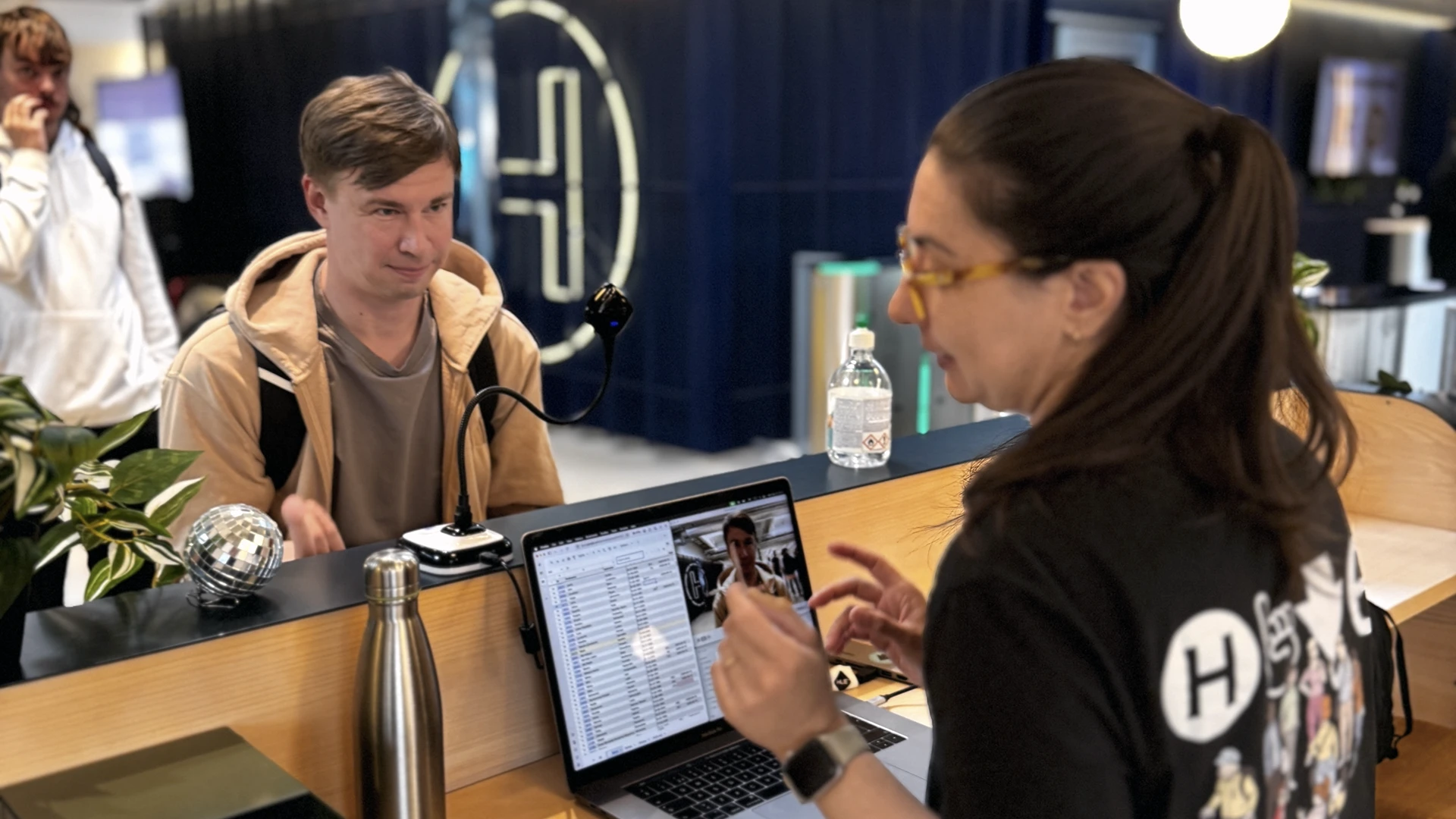 Reception desk scene: a staff member with dark hair in a ponytail and yellow-framed glasses gestures at a laptop screen showing a photo while speaking to a man seated across the counter; a stainless water bottle, potted plant and a plastic water bottle sit on the desk and a blue wall with a circular logo is visible in the background.