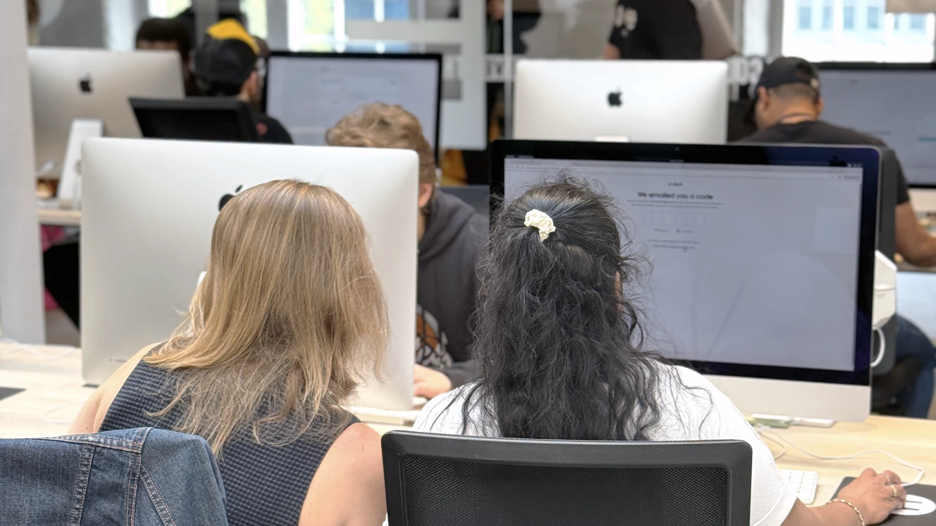 Two people seen from behind sitting side-by-side at large desktop computers in a bright computer lab, one with blonde hair and one with dark hair tied back, while others work at rows of monitors in the background.