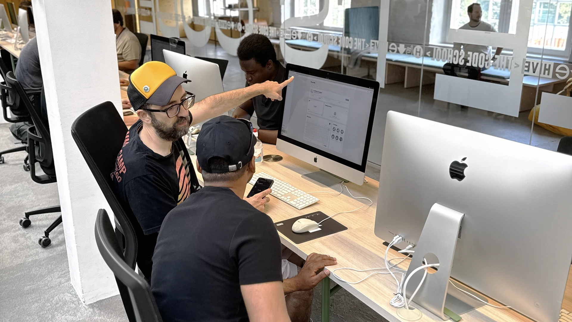 Two coworkers sit at adjacent iMacs in a modern open-plan office — a bearded man wearing a yellow cap and glasses points at one monitor while a colleague in a black cap looks on holding a phone, with other workers and desks visible in the background.