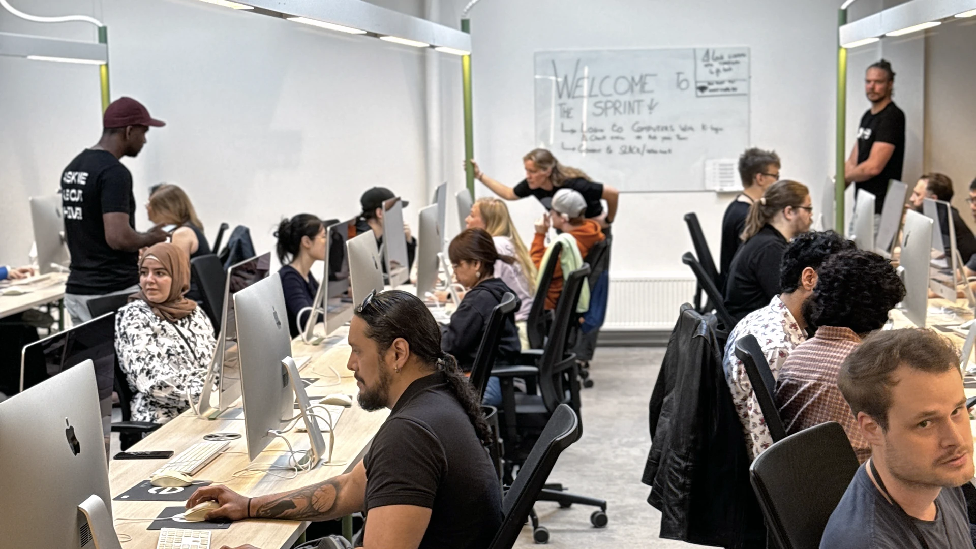 Wide shot of a busy open-plan computer classroom or office with people seated in rows at desktop Macs, focused on their screens while a few staff stand near a whiteboard at the back that has "Welcome" written on it.