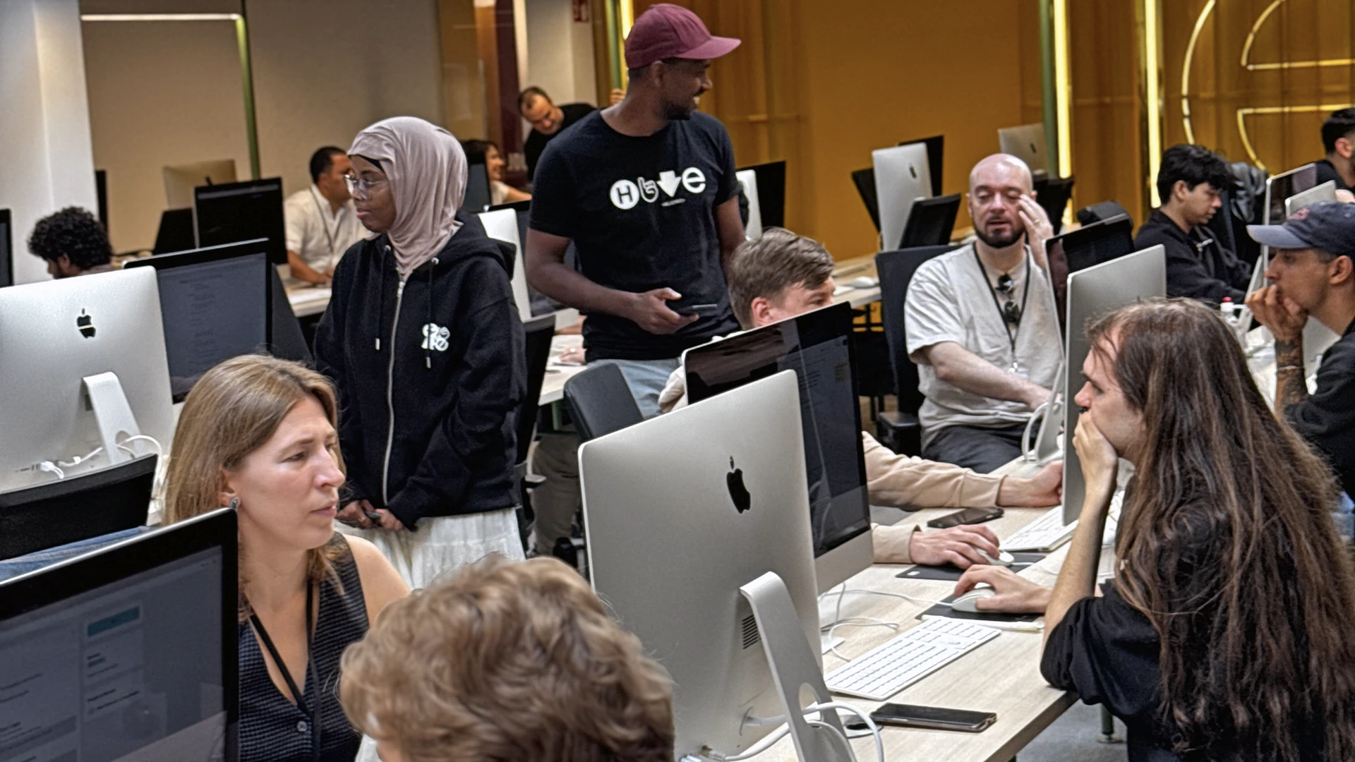 Busy open-plan computer lab with people seated at rows of iMacs and others standing, talking and collaborating; visible individuals include a person wearing a headscarf and another wearing a baseball cap.