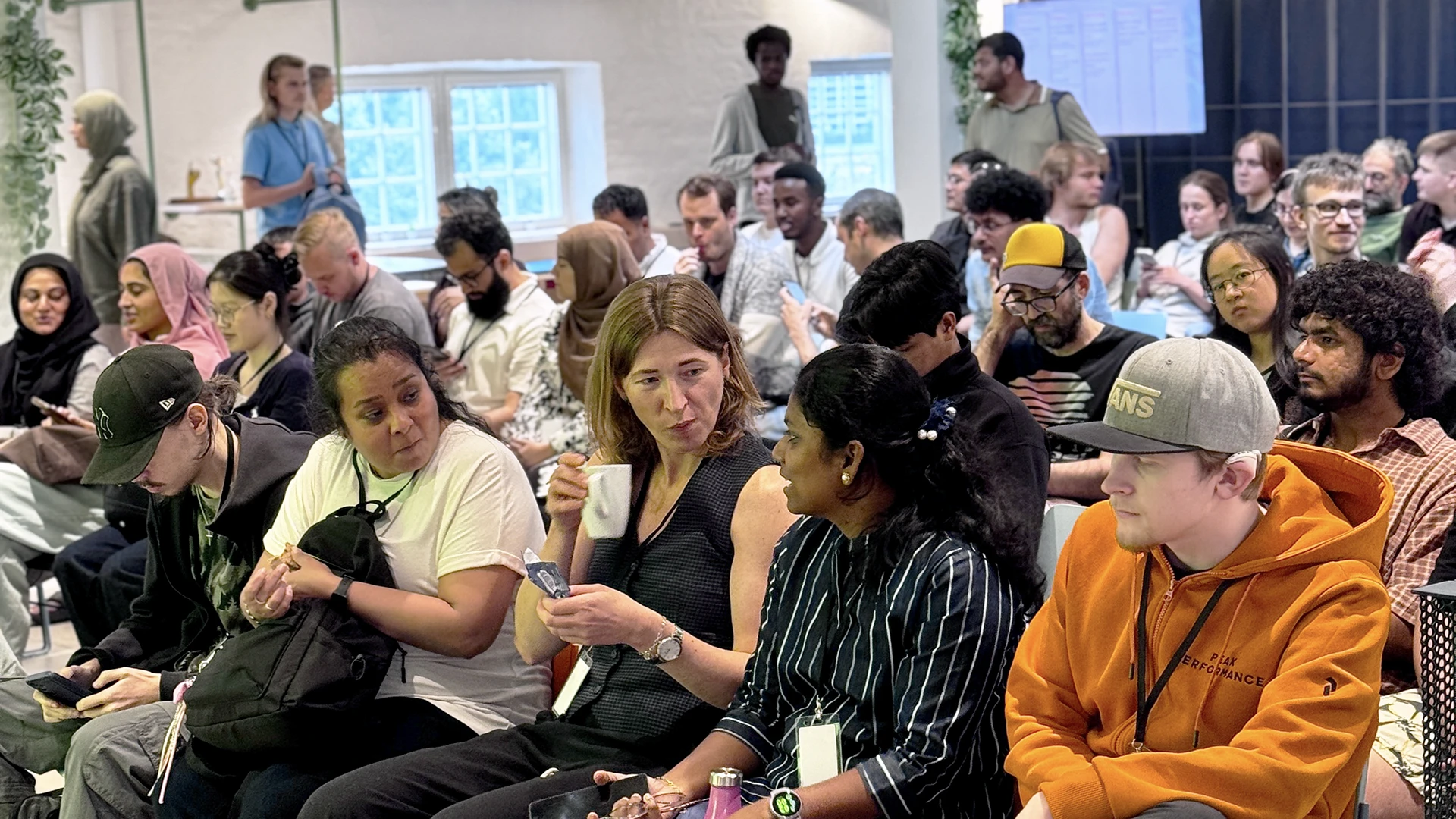 Crowded indoor meeting room with adults seated in rows, many talking or checking phones, a woman near the center holding a cup, a person in an orange hoodie at the right, large windows and a presentation area in the background