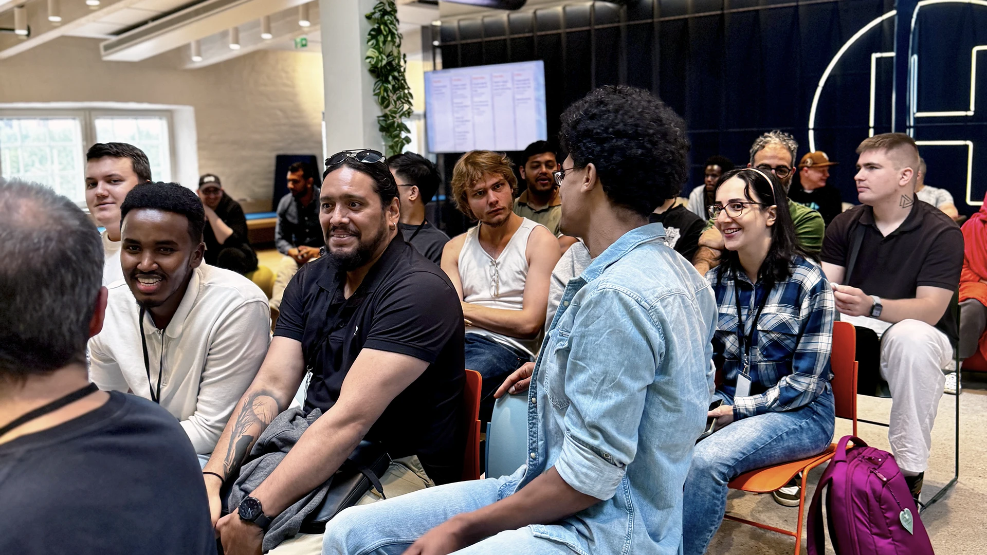 A diverse group of young adults seated in rows in a bright, modern event space, smiling and chatting while facing a presentation area with a large screen and wall logo, with casual clothing, backpacks and plants visible.