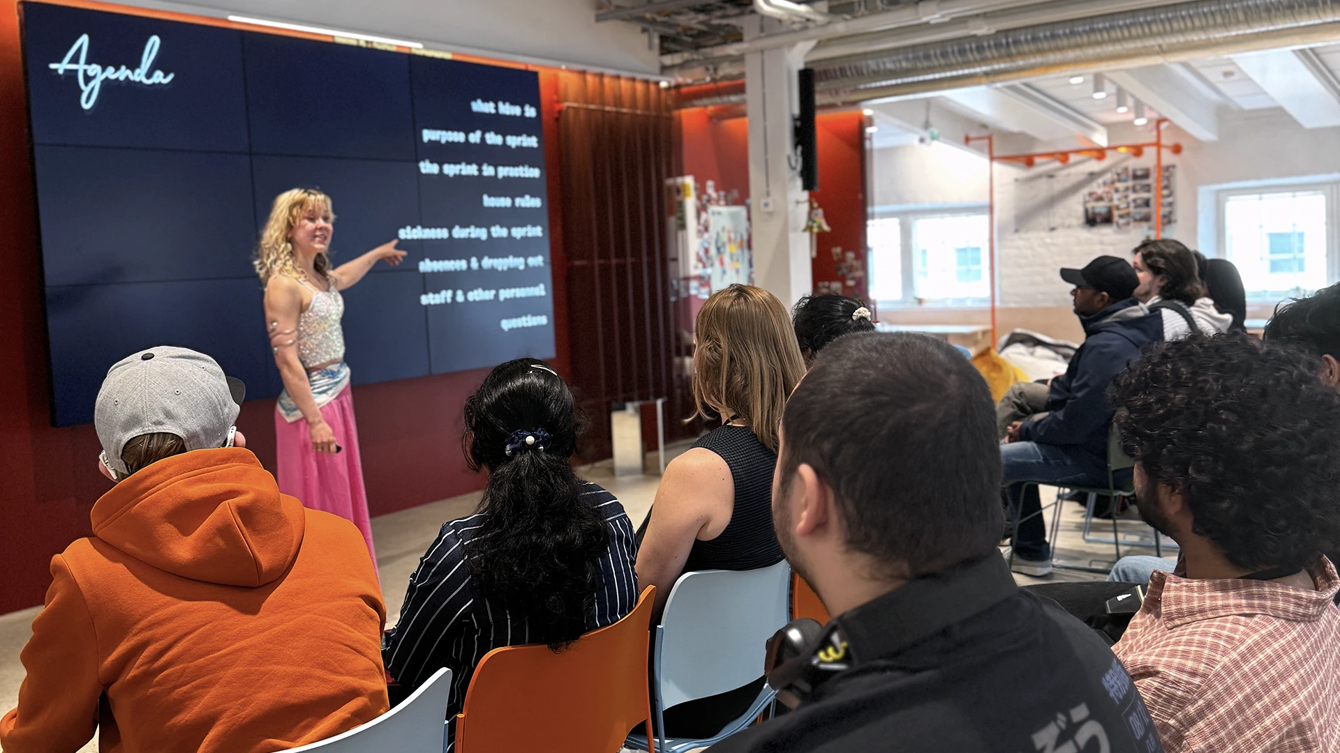 Presenter standing next to a large wall-mounted screen displaying the word "Agenda" and bulleted text, pointing to it while a seated audience in colorful chairs watches in a modern open-office meeting room with exposed ceiling and windows.